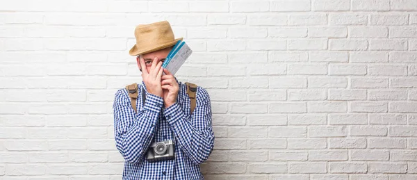 Young traveler man wearing backpack and a vintage camera feels worried and scared, looking and covering face, concept of fear and anxiety. Holding a boarding pass.