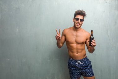 Bir grunge karşı bir mayo giyen genç atletik erkek duvar eğlenceli ve mutlu, pozitif ve doğal, zafer, barış kavramı bir jesti yapıyor