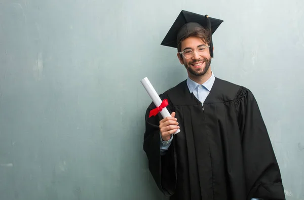 Young graduated man against a grunge wall with a copy space cheerful and with a big smile, confident, friendly and sincere, expressing positivity and success
