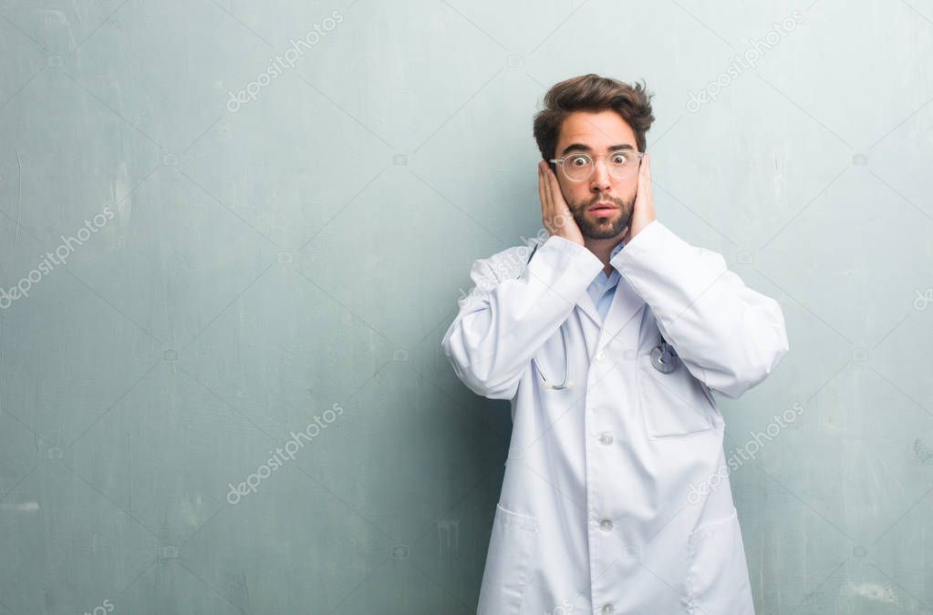 Young friendly doctor man against a grunge wall with a copy space covering ears with hands, angry and tired of hearing some sound