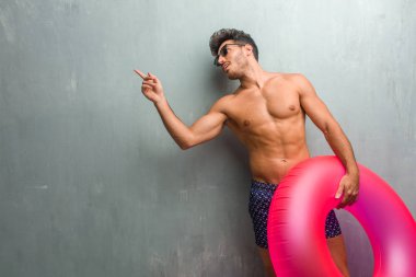 Sürpriz bir şey, doğal ve rahat sunan gülümseyen tarafına işaret eden bir grunge duvara bir mayo giyen genç atletik adam