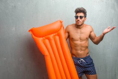 Bir grunge duvar şüphe karşı bir mayo giyiyor ve shrugging genç atletik erkek omuzlar, kararsızlık ve güvensizlik, hakkında bir şey belirsiz kavram