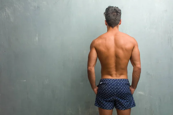 Young athletic man wearing a swimsuit against a grunge wall showing back, posing and waiting, looking back