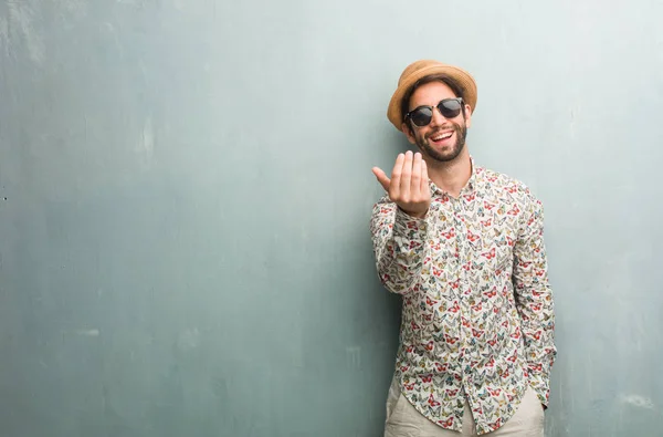 Young traveler man wearing a colorful shirt inviting to come, confident and smiling making a gesture with hand, being positive and friendly