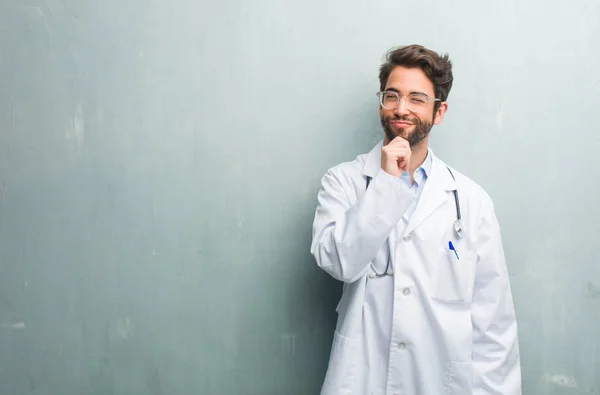 Young friendly doctor man against a grunge wall with a copy space thinking and looking up, confused about an idea, would be trying to find a solution