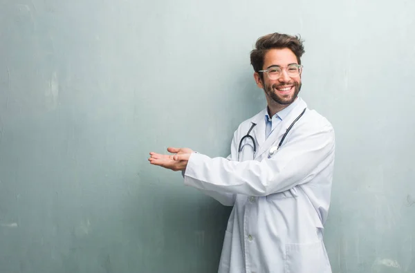 Young friendly doctor man against a grunge wall with a copy space holding something with hands, showing a product, smiling and cheerful, offering an imaginary object