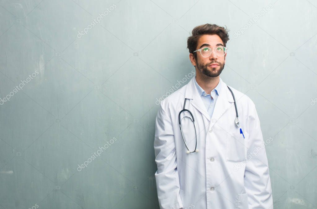 Young friendly doctor man against a grunge wall with a copy space looking up, thinking of something fun and having an idea, concept of imagination, happy and excited