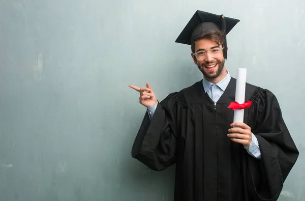 Young graduated man against a grunge wall with a copy space pointing to the side, smiling surprised presenting something, natural and casual