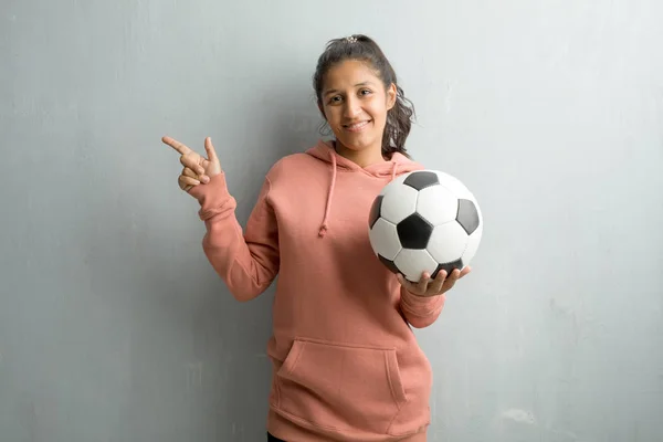 壁側を指している若いスポーティなインド女性は 笑みを浮かべて何か 自然に さりげなく提示を驚かせた ゲームをプレイするサッカー ボールを保持しています — ストック写真