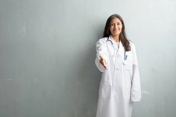 Young indian doctor woman against a wall reaching out to greet someone or gesturing to help, happy and excited