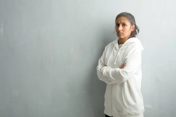 Young sporty indian woman against a gym wall crossing his arms, serious and imposing, feeling confident and showing power