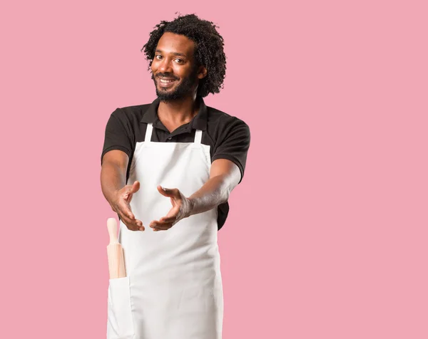 Handsome african american baker reaching out to greet someone or gesturing to help, happy and excited