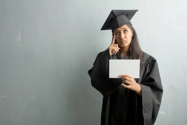 Young graduated indian woman against a wall thinking and looking up, confused about an idea, would be trying to find a solution. Holding a placard.