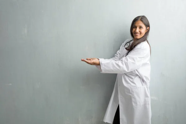 Young indian doctor woman against a wall holding something with hands, showing a product, smiling and cheerful, offering an imaginary object