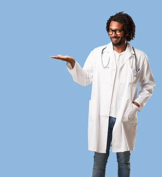 Handsome african american medical doctor holding something with hands, showing a product, smiling and cheerful, offering an imaginary object