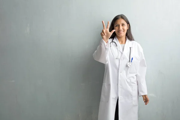 Young indian doctor woman against a wall showing number three, symbol of counting, concept of mathematics, confident and cheerful