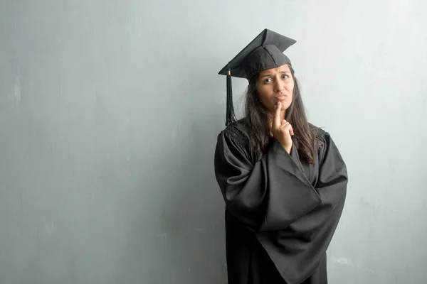 Young graduated indian woman against a wall doubting and confused, thinking of an idea or worried about something