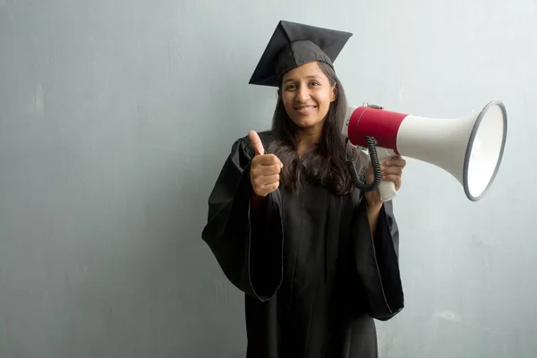 Young graduated indian woman against a wall cheerful and excited, smiling and raising her thumb up, concept of success and approval, ok gesture. Holding a megaphone.