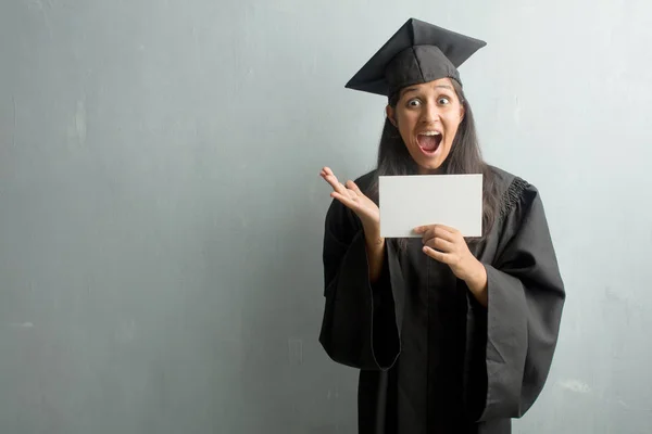 Young graduated indian woman against a wall surprised and shocked, looking with wide eyes, excited by an offer or by a new job, win concept. Holding a placard.