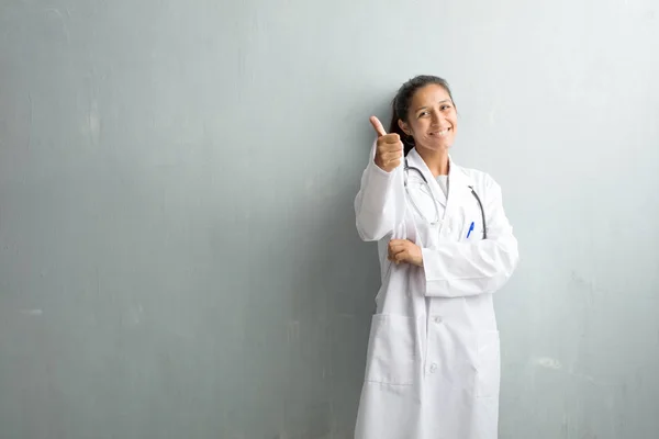 Young indian doctor woman against a wall cheerful and excited, smiling and raising her thumb up, concept of success and approval, ok gesture