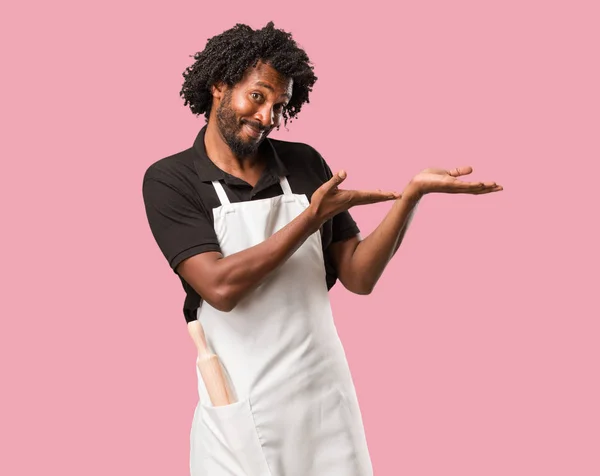 Handsome african american baker holding something with hands, showing a product, smiling and cheerful, offering an imaginary object