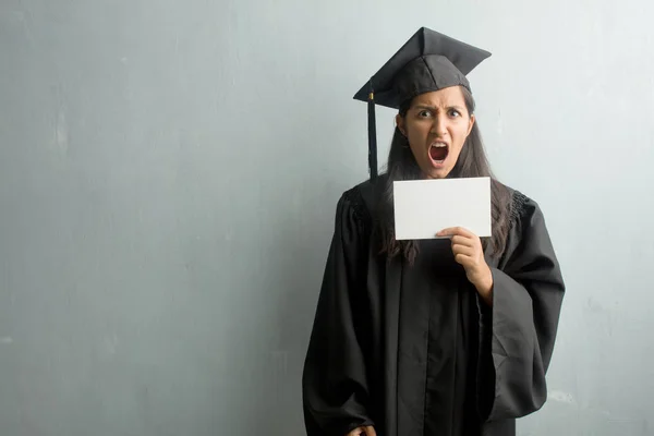 Young graduated indian woman against a wall screaming angry, expression of madness and mental instability, open mouth and half-opened eyes, madness concept. Holding a placard.