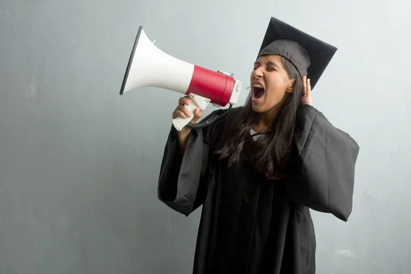 Young graduated indian woman against a wall crazy and desperate, screaming out of control, funny lunatic expressing freedom and wild. Holding a megaphone.