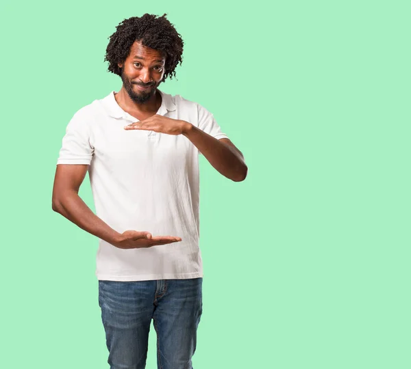 Handsome african american holding something with hands, showing a product, smiling and cheerful, offering an imaginary object