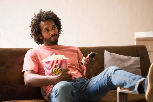 Young african man watching television, he is bored, holding a popcorn bucket.