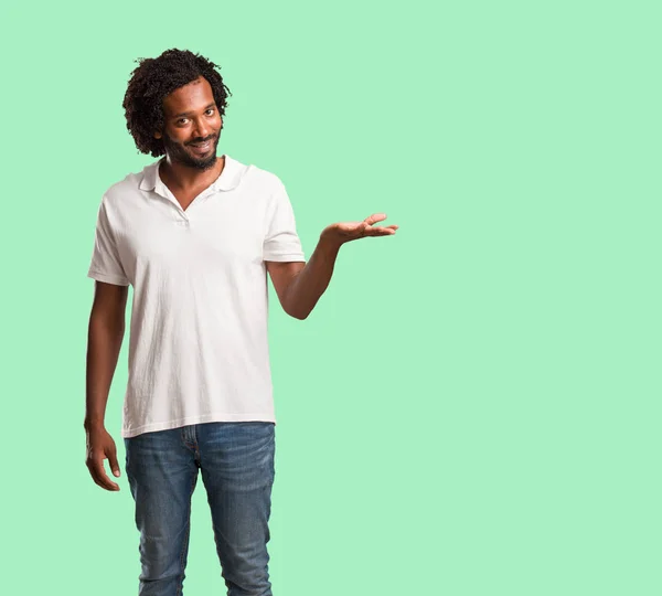 Handsome african american holding something with hands, showing a product, smiling and cheerful, offering an imaginary object