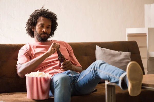 Young african man watching a movie holding a popcorn bucket and drinking a beer.