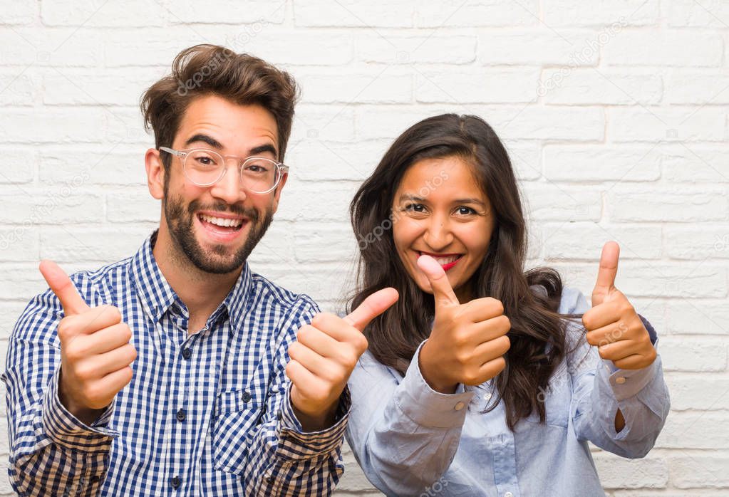 Young indian woman and caucasian man couple cheerful and excited, smiling and raising her thumb up, concept of success and approval, ok gesture