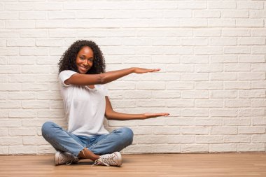 Duvara karşı bir ürün gösterilen bir ahşap zemin üzerinde oturan genç siyah kadın