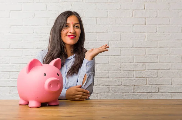 Young indian woman holding something with hands, showing a product, smiling and cheerful, offering an imaginary object. Holding a piggy bank.