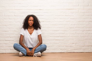 Young black woman sitting on a wooden floor doubting and confused against brick wall
