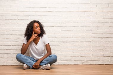 Young black woman sitting on a wooden floor doubting and confused against brick wall