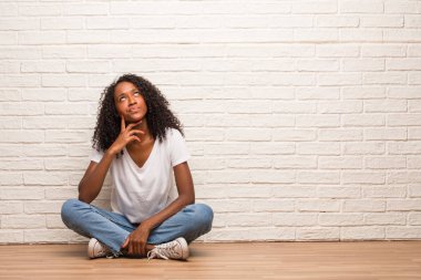 Young black woman sitting on a wooden floor doubting and confused against brick wall