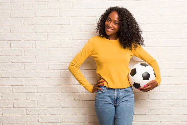 Young black woman with soccer ball with hands on hips against brick wall