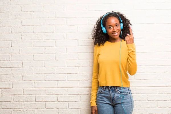 Young black woman in headphones showing number one against brick wall