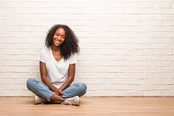 陽気で笑顔のレンガの壁に木の床の上に座って若い黒人女性 — ストック写真
