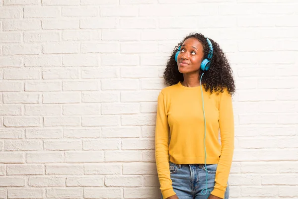 Young black woman in headphones looking up, thinking of something fun against brick wall