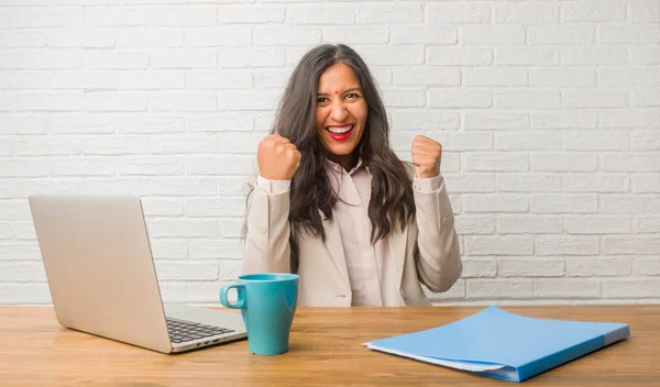 Young indian woman at office very happy and excited, raising arms, celebrating victory or success