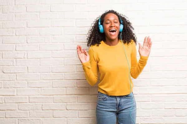 Young black woman in headphones laughing and having fun, rising arms against brick wall