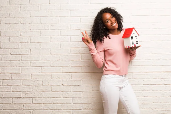 Young black woman with model house making gesture of victory against brick wall