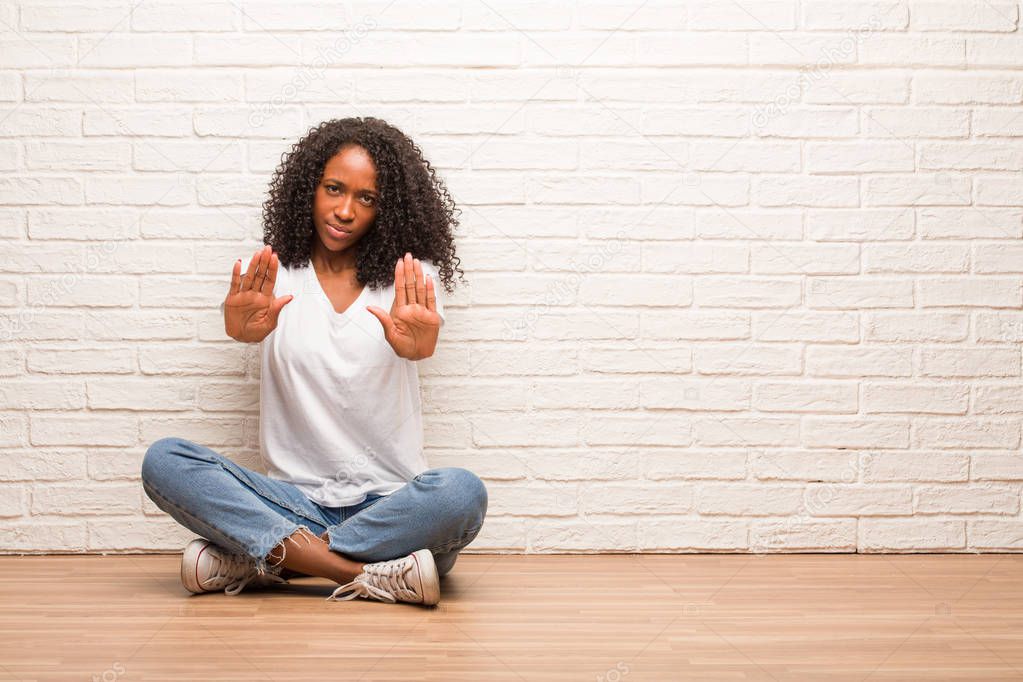 Young black woman sitting on wooden floor serious and determined, putting hand in front, stop gesture against brick wall