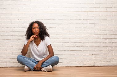 Young black woman sitting on a wooden floor doubting and confused against brick wall