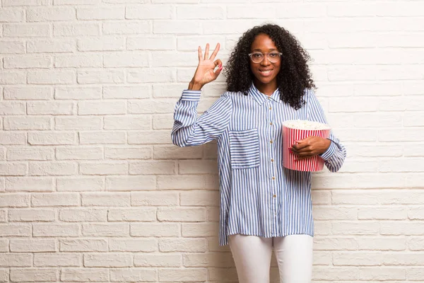 年轻的黑人妇女与爆米花桶开朗和自信做 手势反对砖墙 — 图库照片