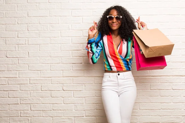 Young black woman with shopping bags crossing fingers against brick wall