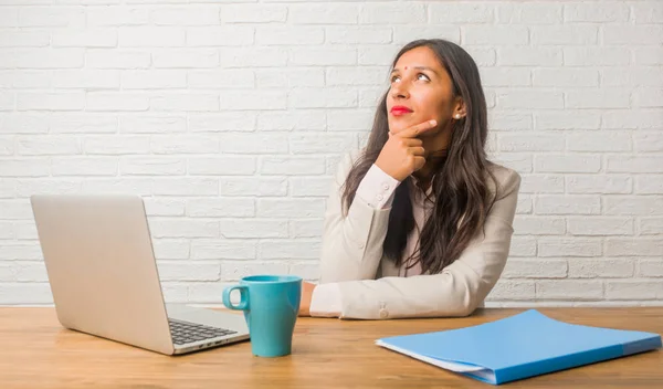 Young indian woman at office thinking and looking up, confused about an idea, would be trying to find solution