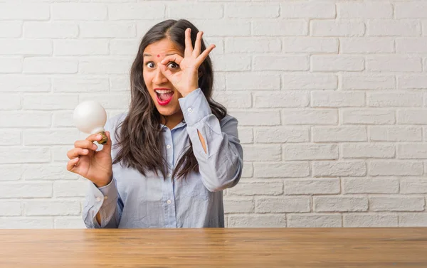 Young indian woman cheerful and confident doing ok gesture, excited and screaming, concept of approval and success. Holding a light bulb.